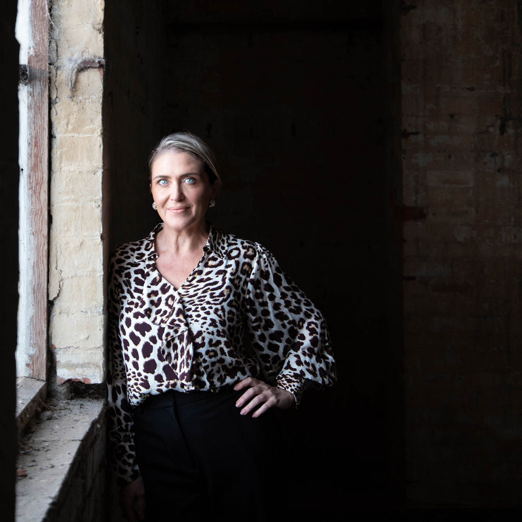 Felicity wearing a leopard print blouse standing against a dark wall with a window on the left.