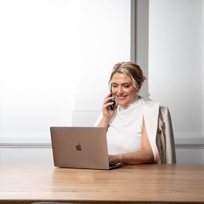 A woman in a white sleeveless top sits at a wooden desk, talking on a mobile phone with an open silver laptop in front of her. The background is a bright, modern room with white walls and paneling.