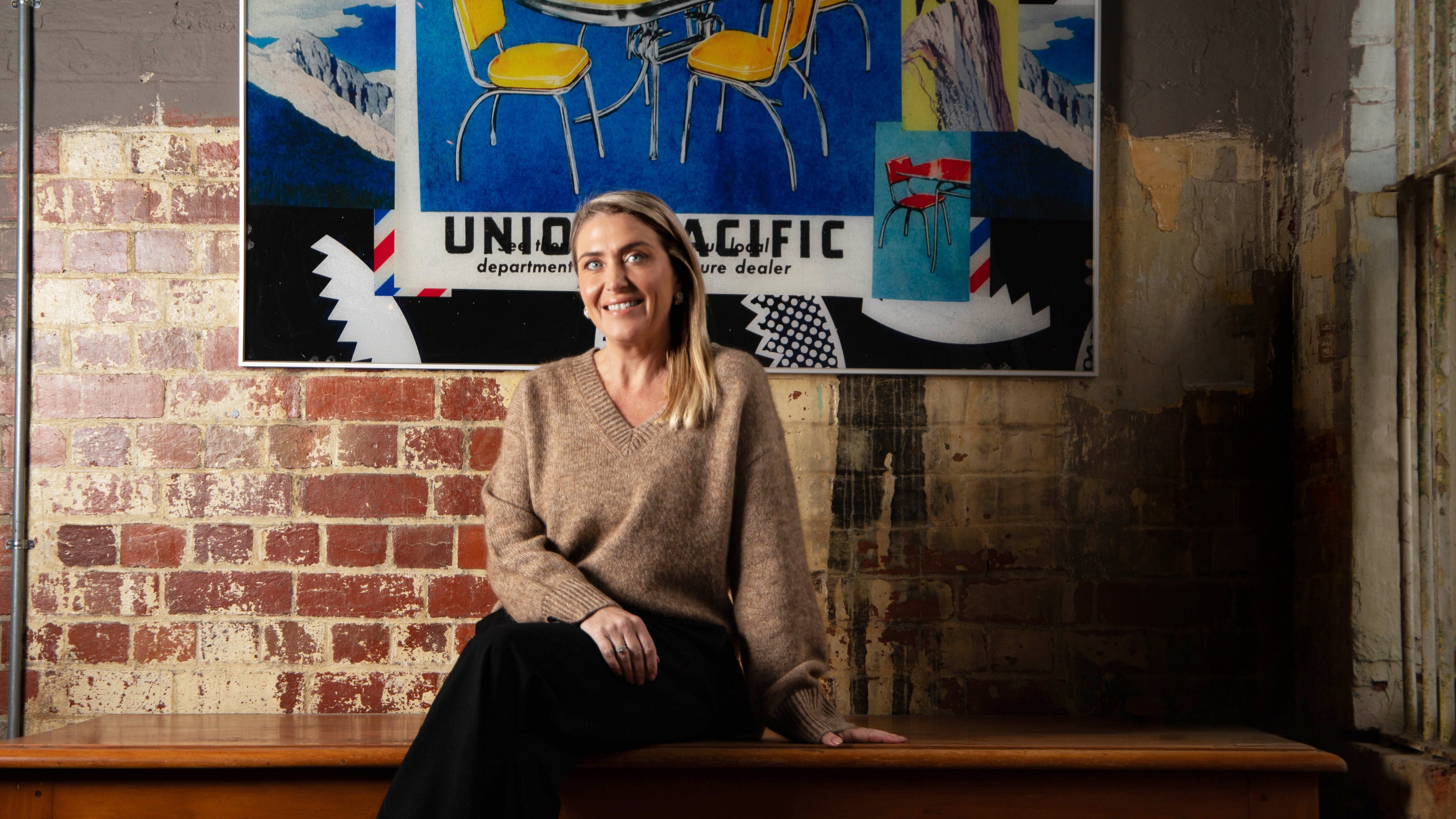 Woman sitting on a wooden bench in front of a brick wall with a colorful poster above.