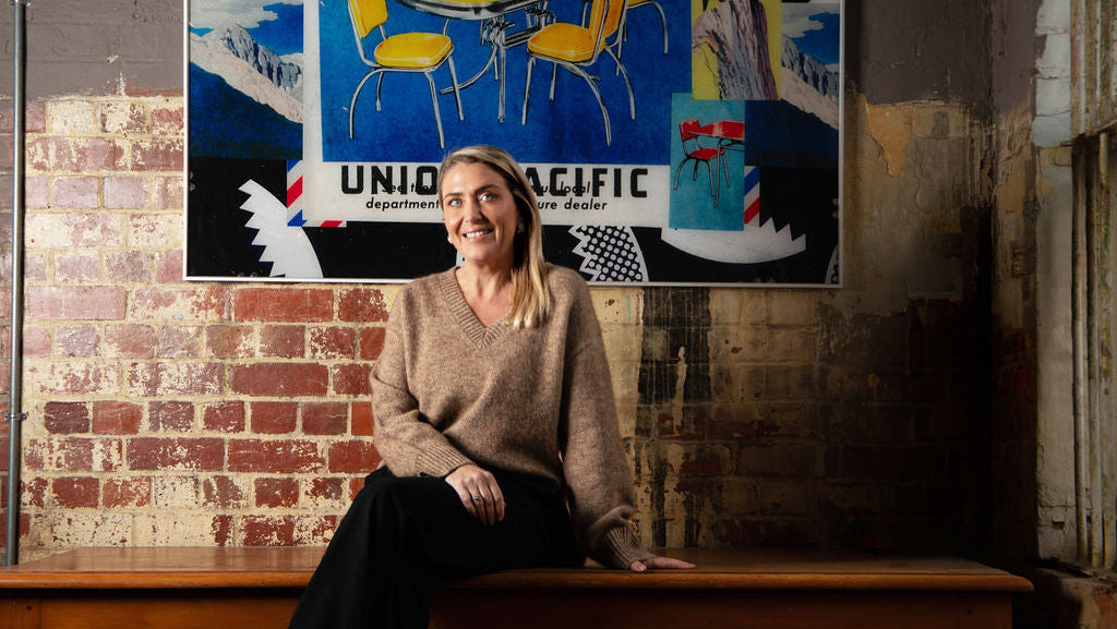 A woman with blonde hair sitting on a wooden bench in front of a brick wall, wearing a beige knit sweater and black pants. A large vintage-style Union Pacific poster hangs on the wall behind her.