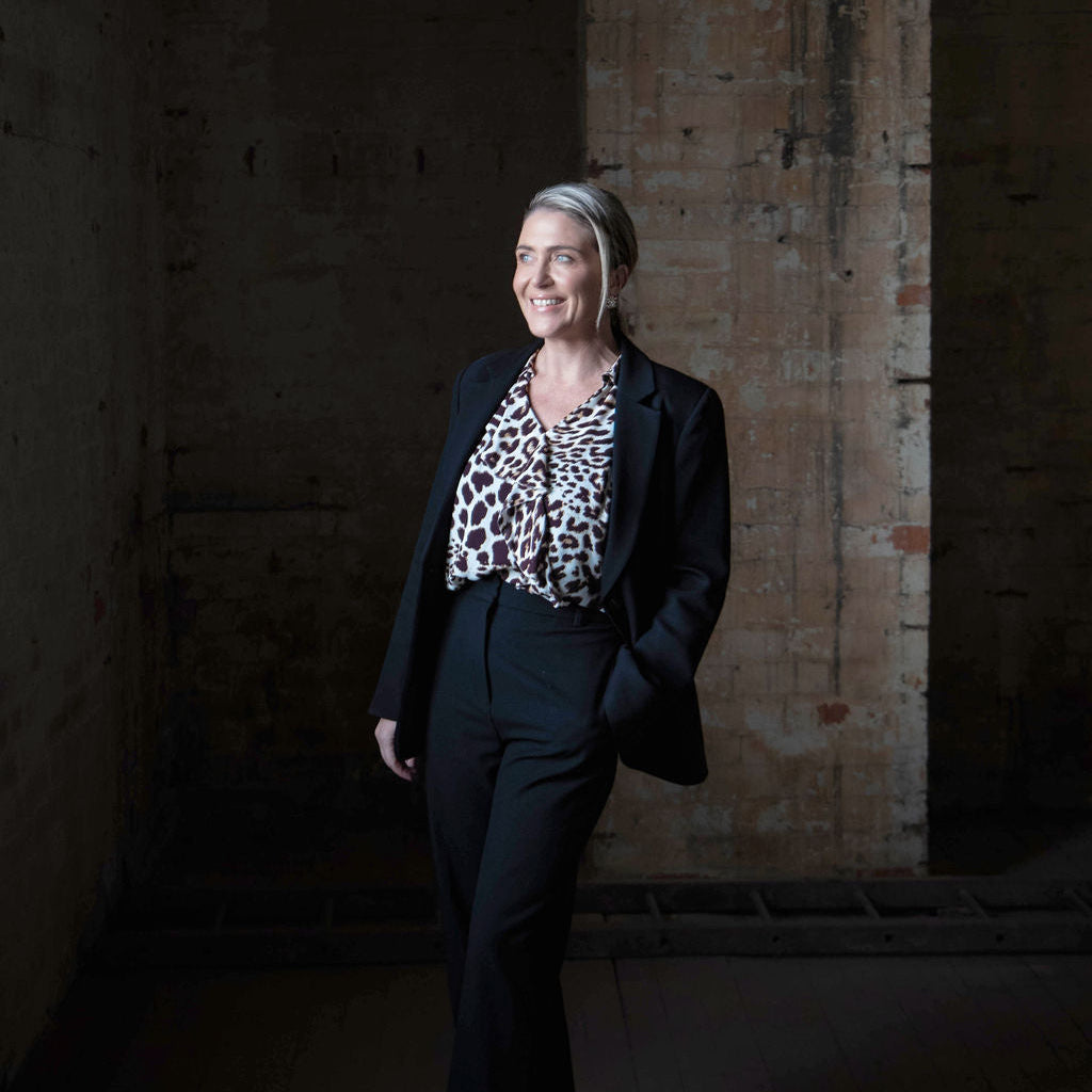 Woman in a dark suit with a patterned top standing against a textured wall.