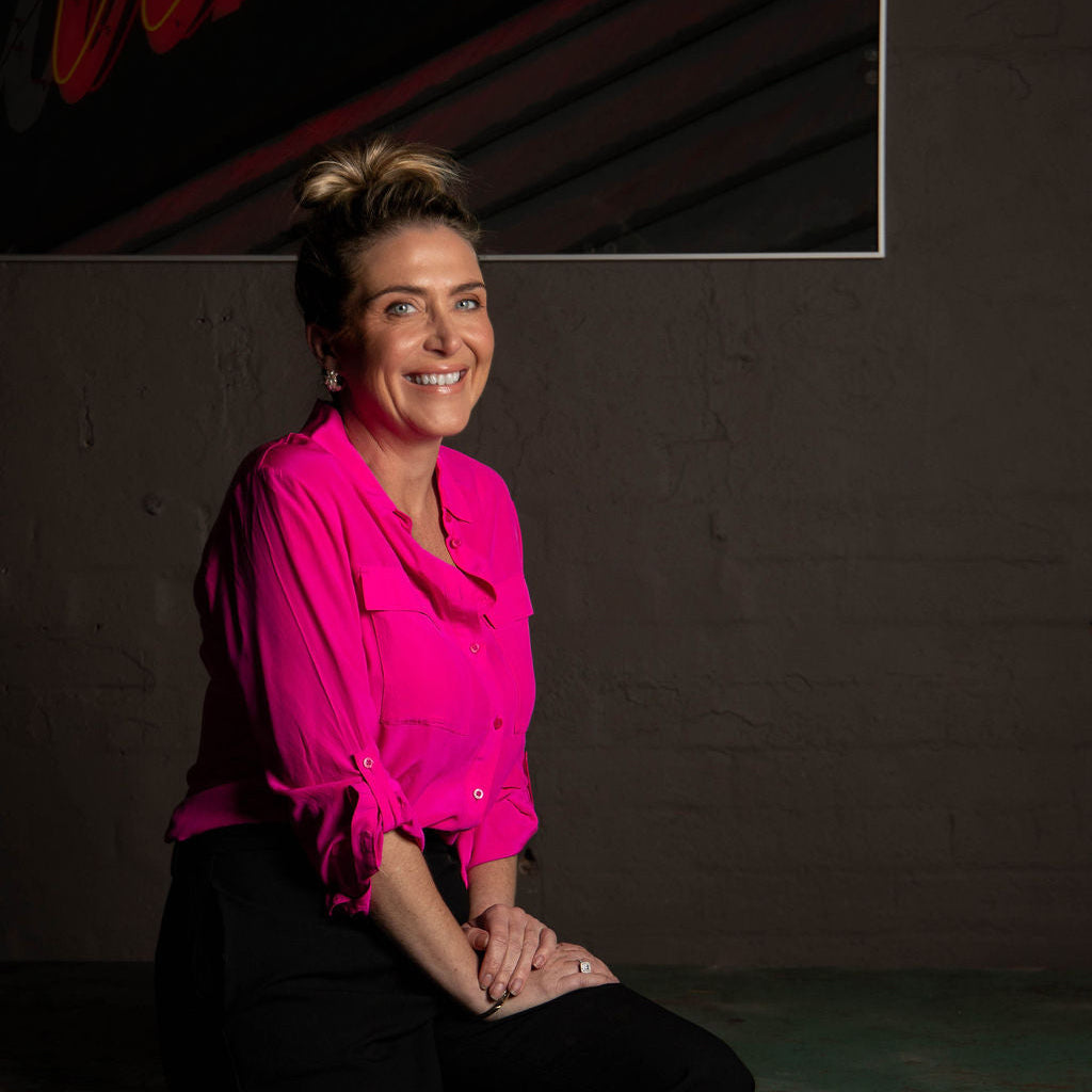 Woman in a bright pink blouse sitting against a dark wall with a neon sign in the background.