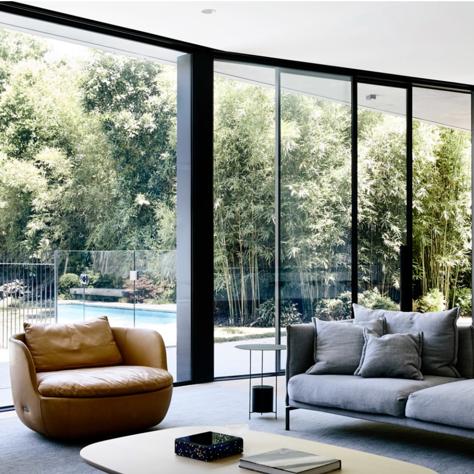 A modern living room interior with large floor-to-ceiling glass windows, black metal frames, outdoor greenery, a pool area, a brown leather armchair, a gray sectional sofa, and a white coffee table.