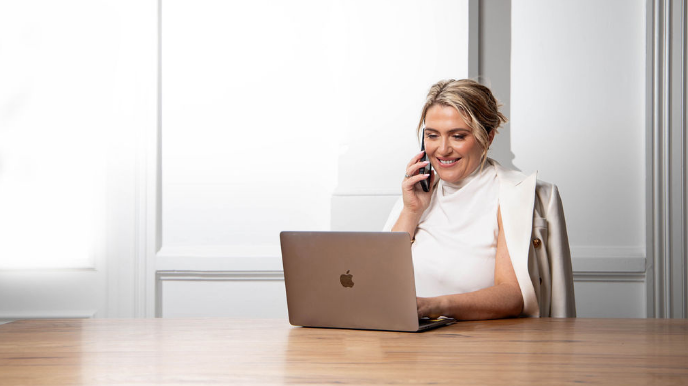 Woman sitting at a desk with a laptop and phone, smiling.