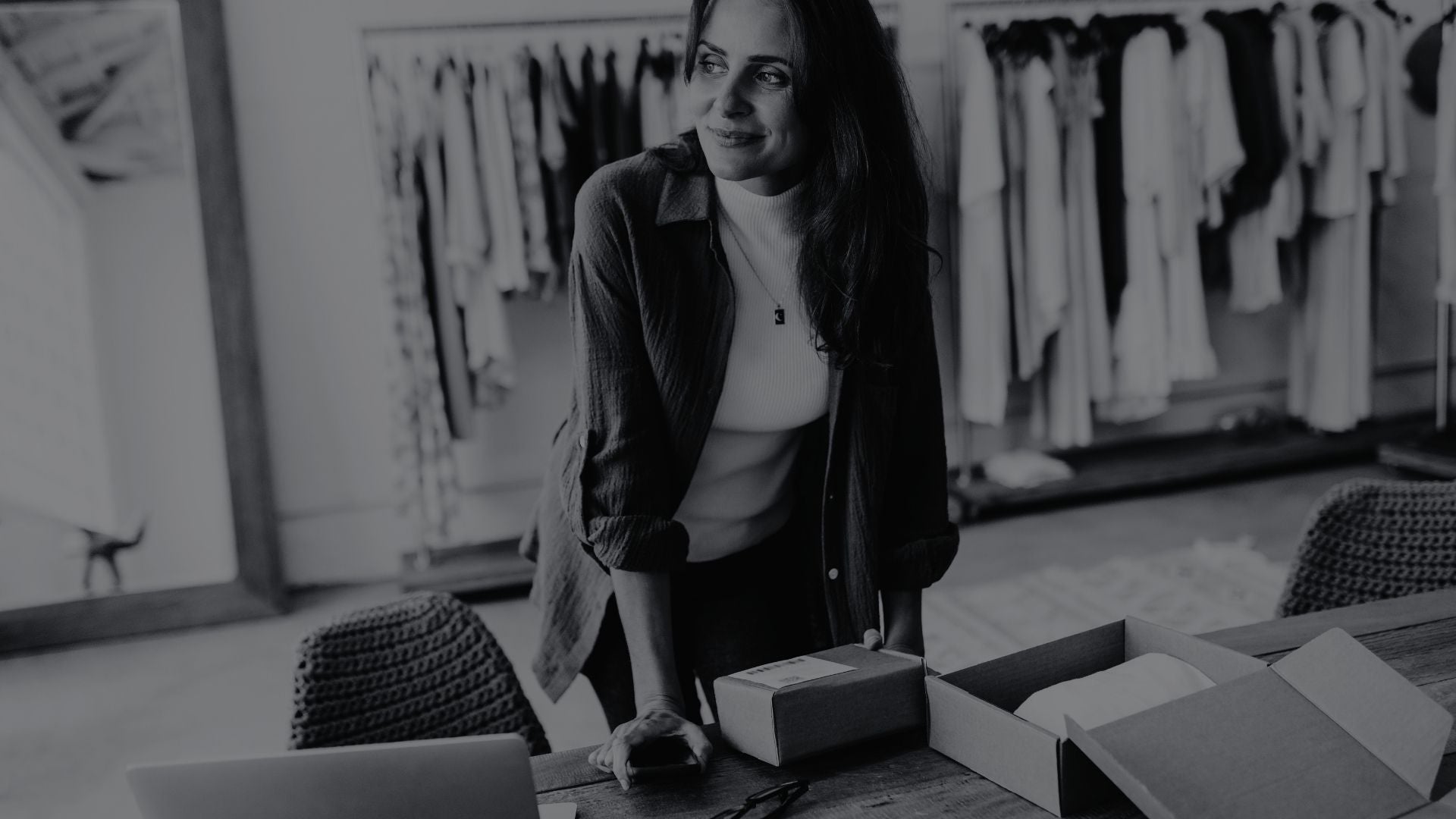 Woman standing in a clothing store with boxes and a laptop on a table.
