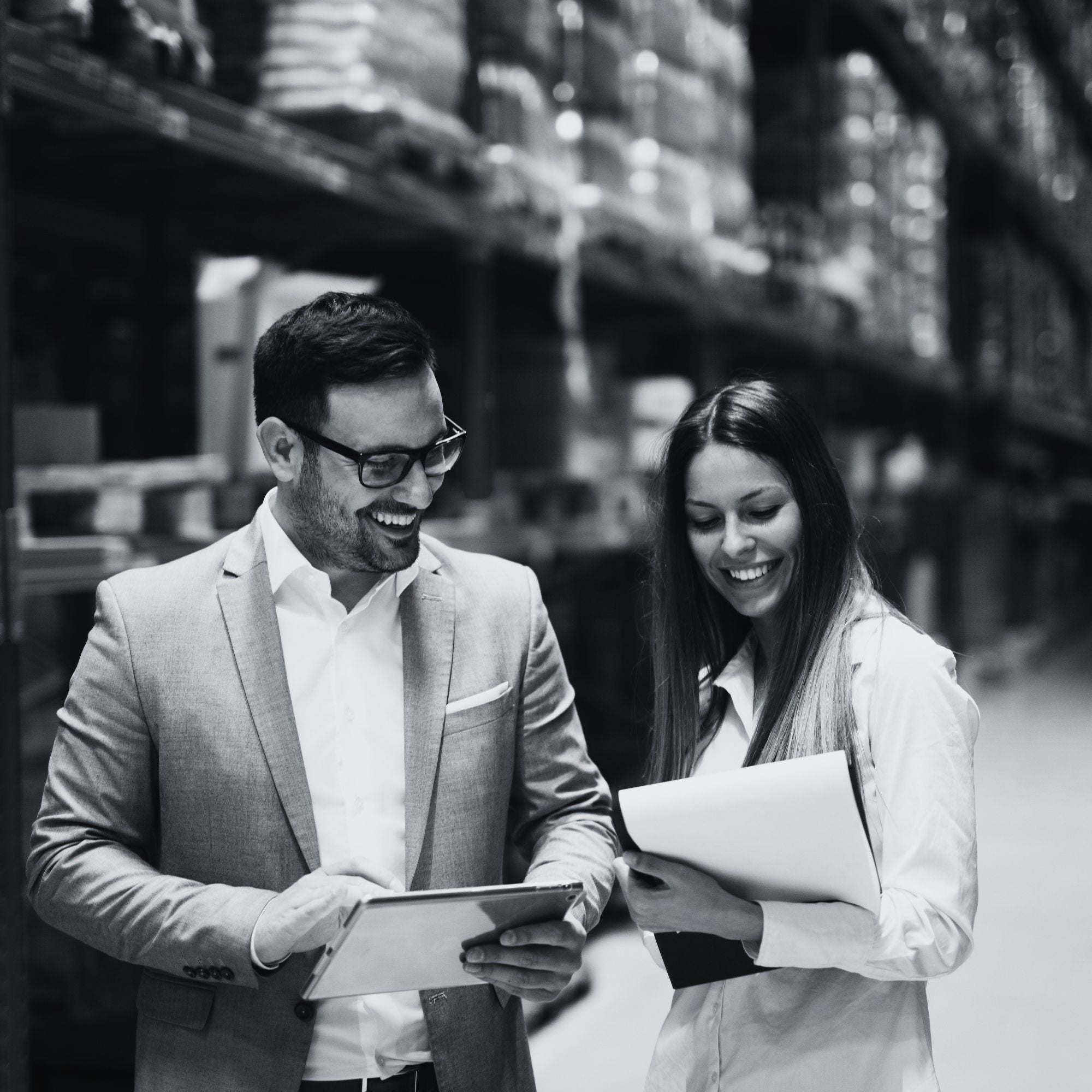 Two people in a warehouse setting, one holding a tablet and the other holding papers.