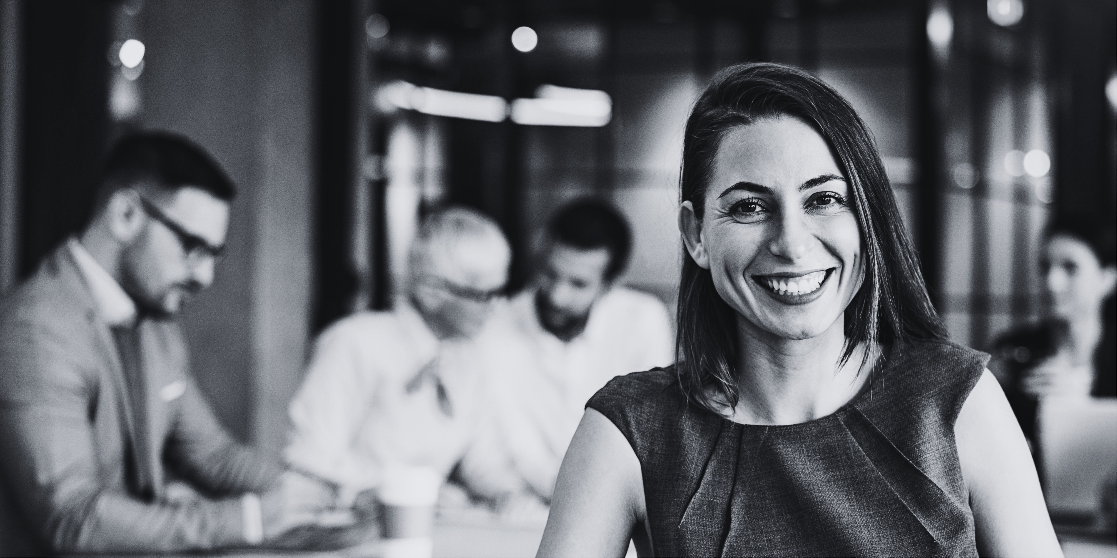 Black and white photo of a woman smiling in an office setting with colleagues in the background.