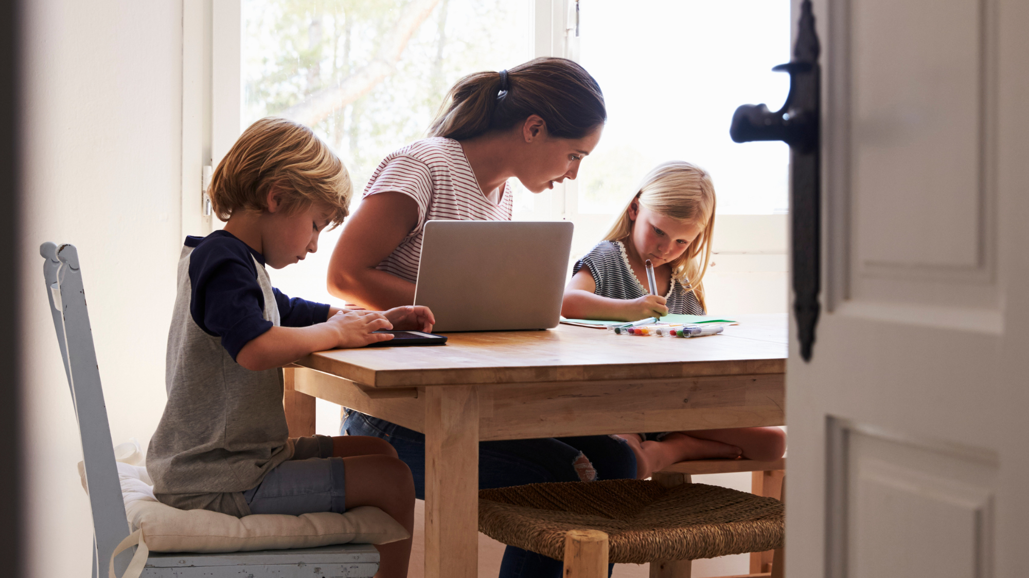 Woman and two children sitting at a table with a laptop and books, in a bright room.